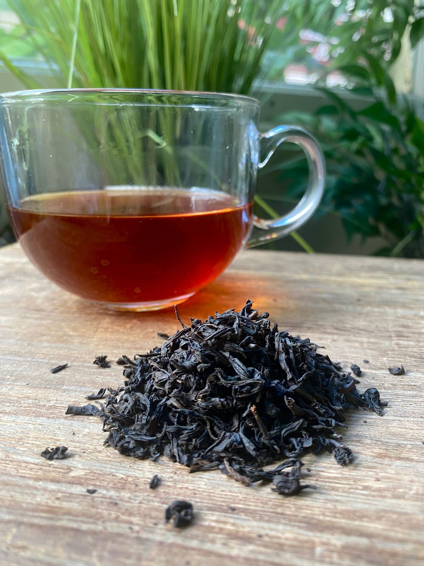 Clear glass mug with black tea and a pile of black tea leaves on a wooden surface with plants in the background.