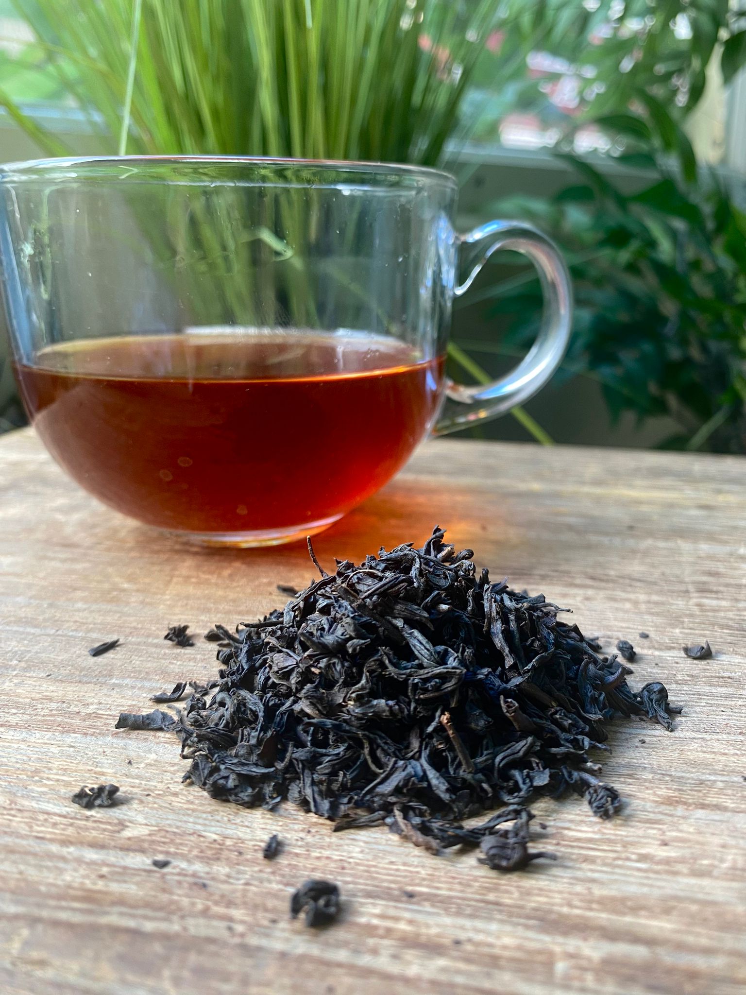 Clear glass mug with black tea and a pile of black tea leaves on a wooden surface with plants in the background.