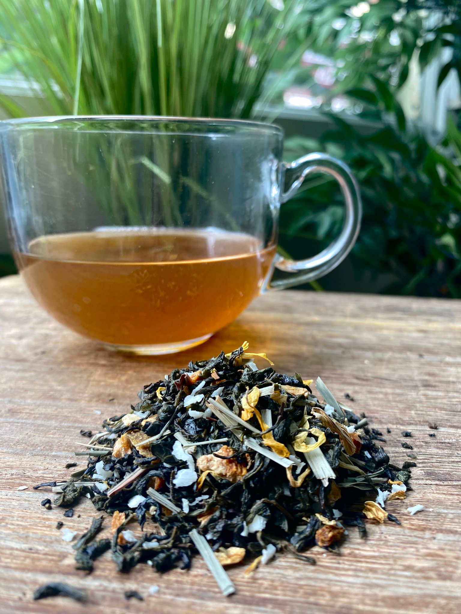 Clear glass mug of lemon meringue tea with a pile of loose leaf tea leaves on a wooden surface with greenery in the background