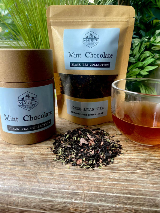 A wooden table displays a package and cylinder of Marie's Mint Chocolate black tea. Loose tea leaves and a glass cup of brewed tea accompany them. Lush greenery sets a fresh tone.