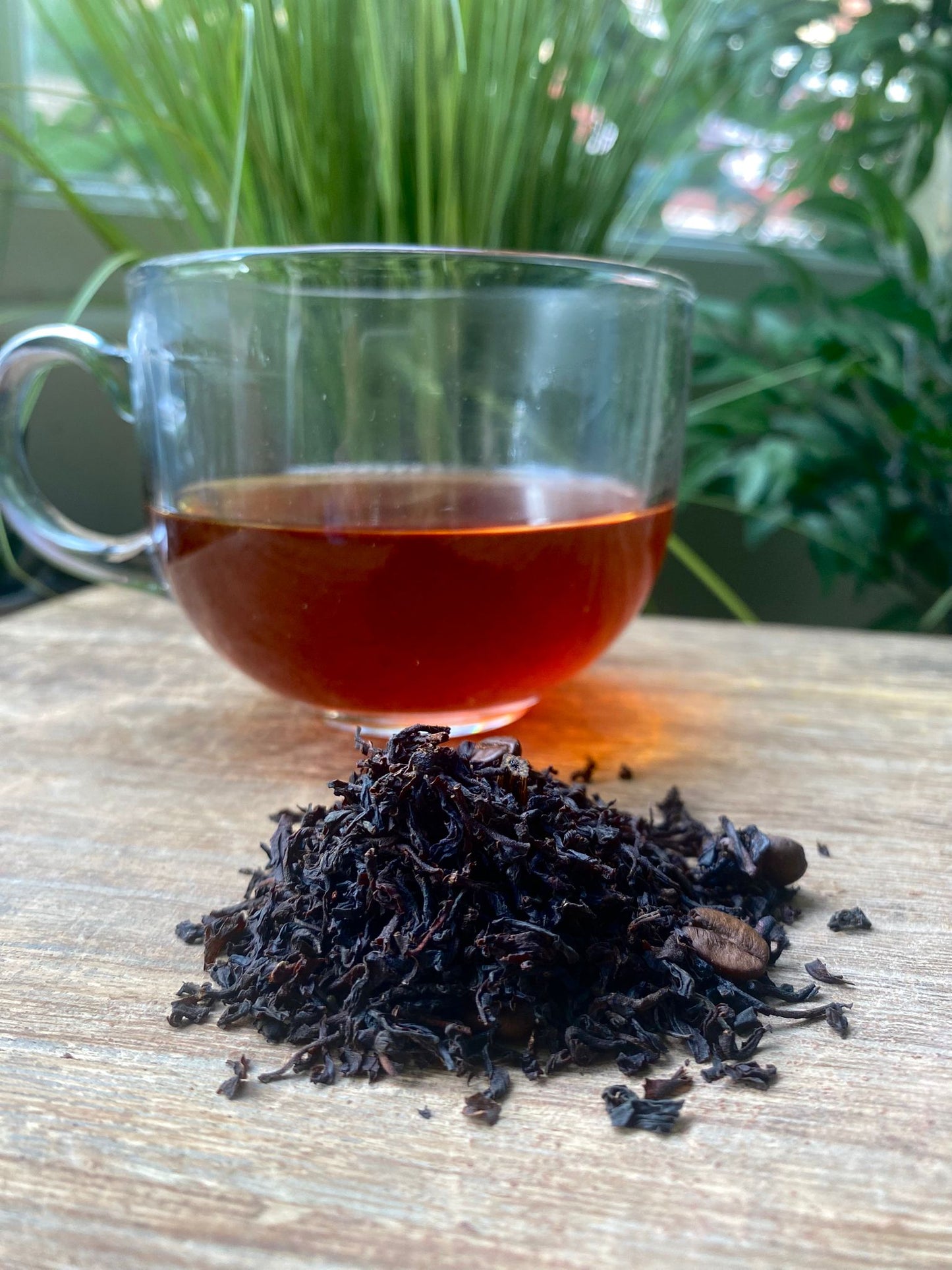 Clear glass mug with Mocha tea and a pile of black tea leaves  with coffee beans scattered throughout on a wooden surface with green plants in the background.