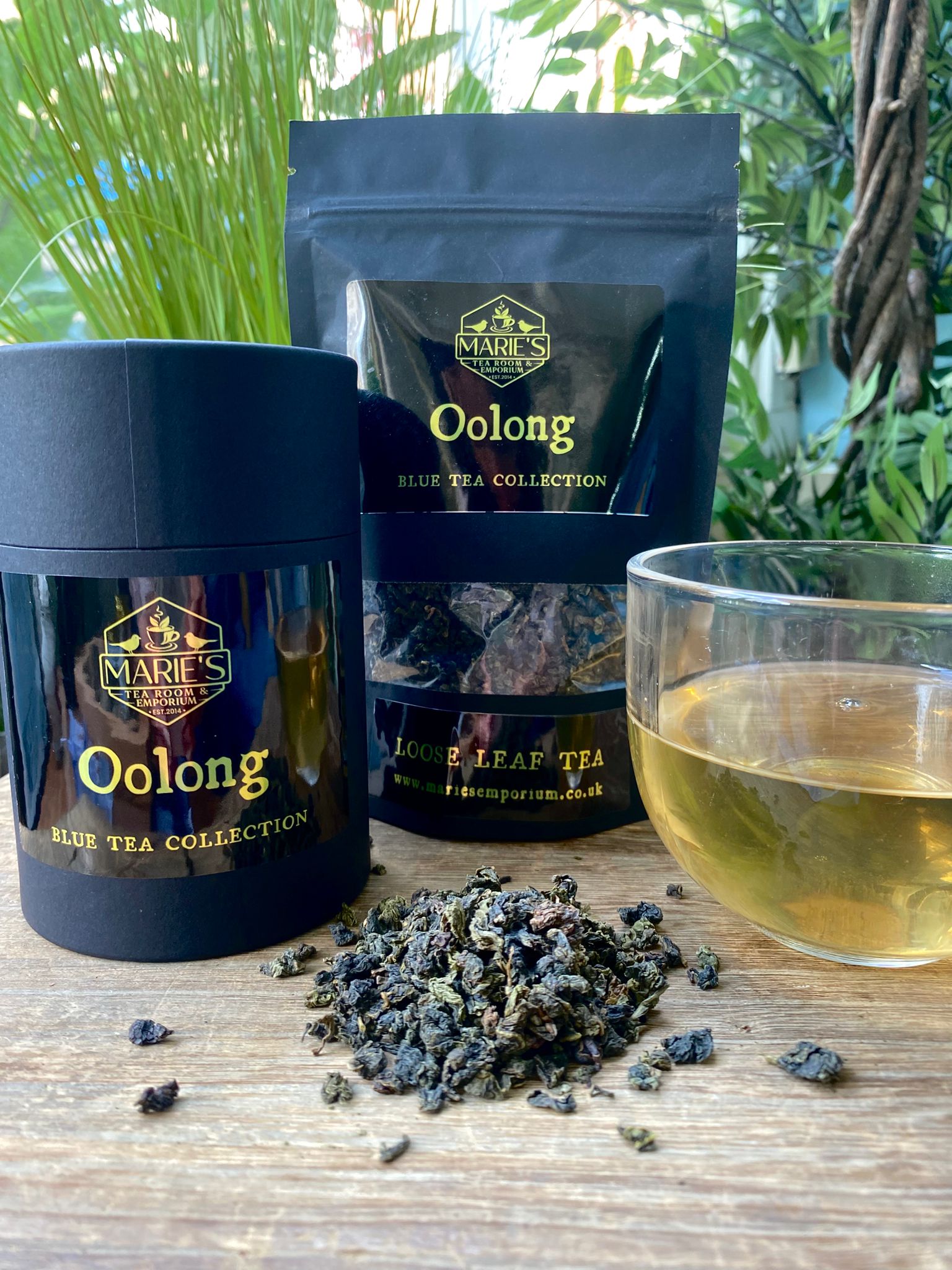 Marie's Blue Tea Collection Oolong tea packaging, loose leaves, and a cup of tea on a wooden surface with greenery in the background.