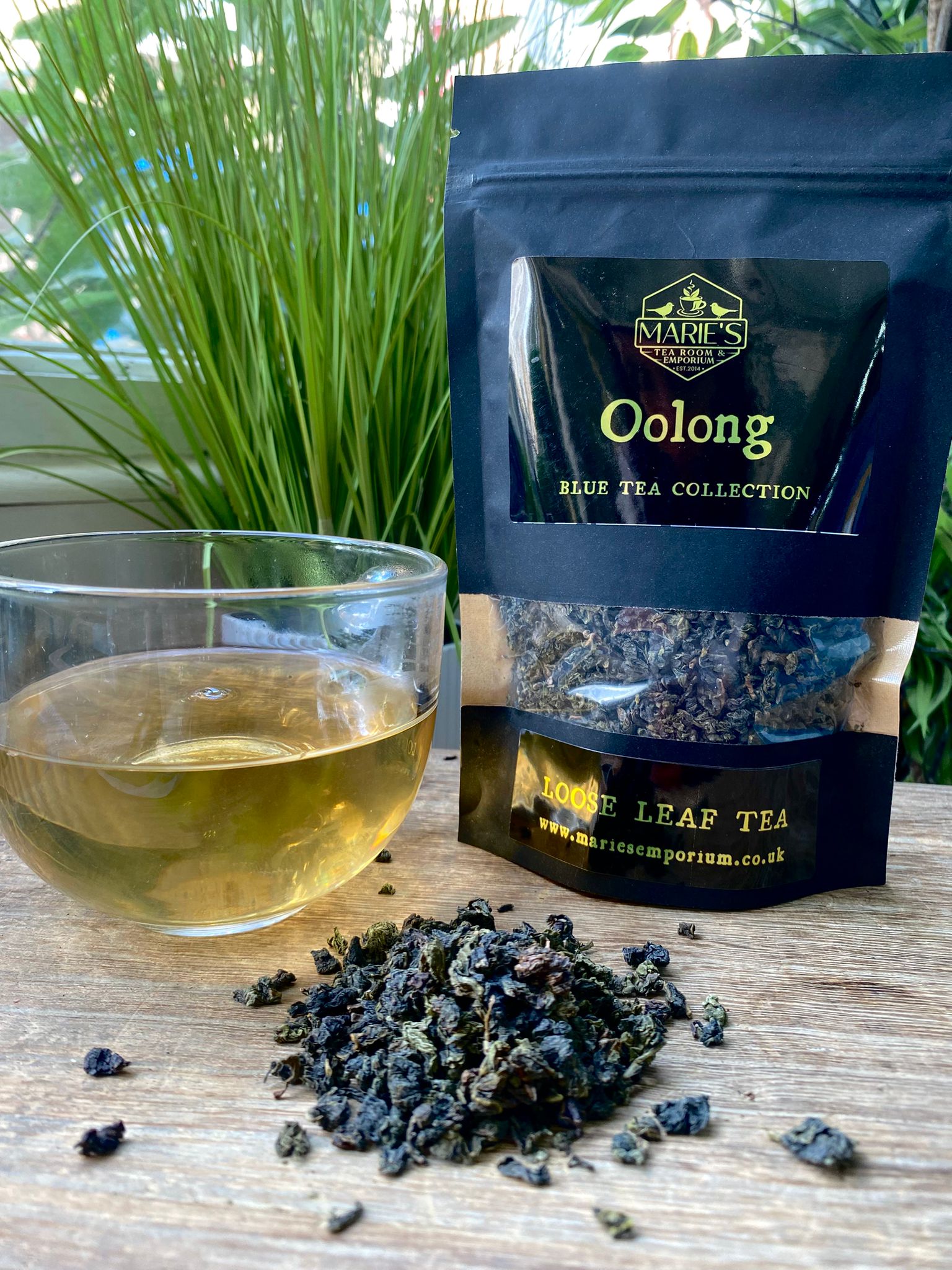 maries Oolong blue tea collection package with a glass of tea and loose tea leaves on a wooden surface.