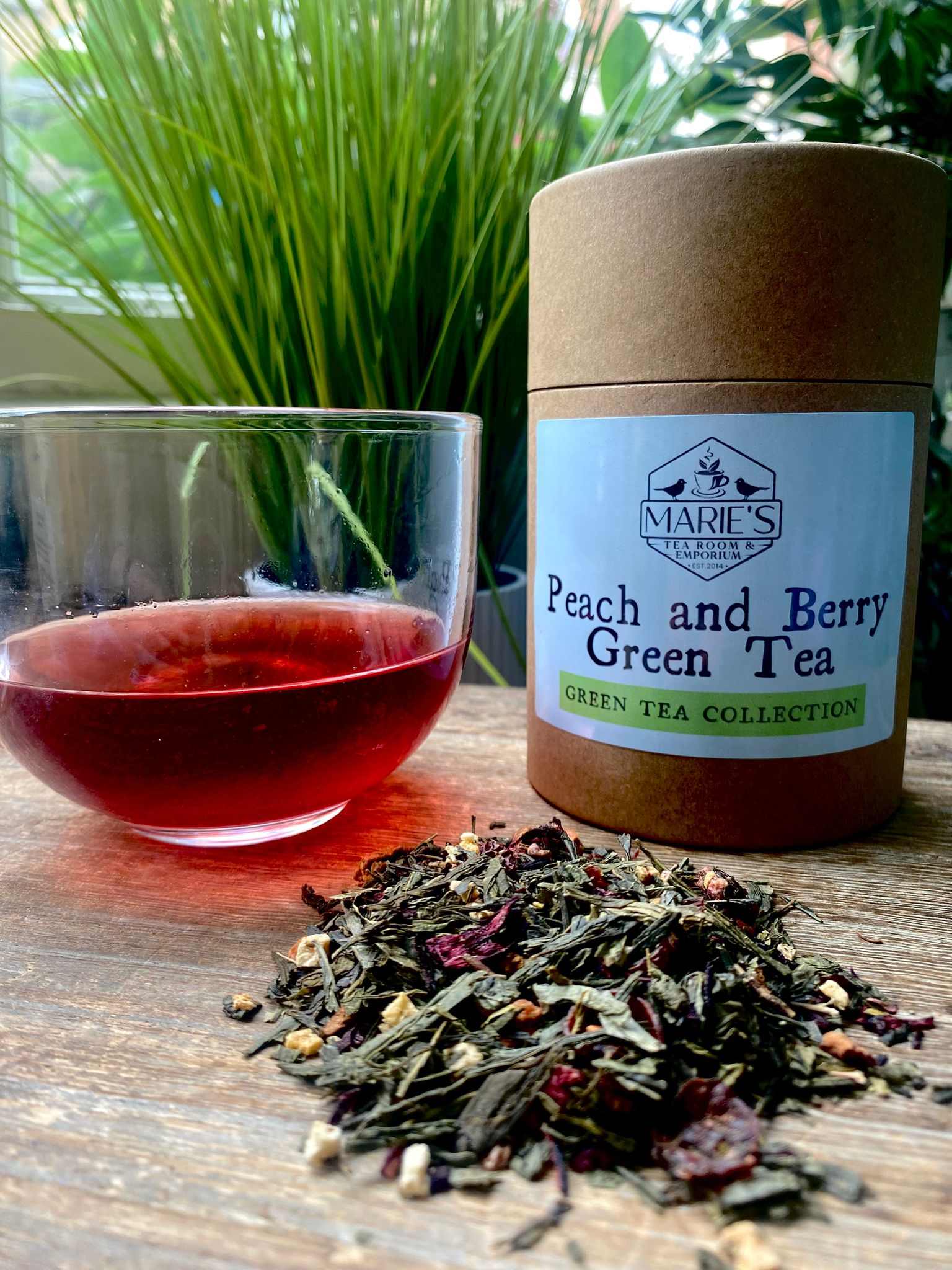 A glass of red tea beside a Marie's Peach and Berry Green Tea container on a wooden table. Loose tea leaves are scattered, with lush greenery behind.