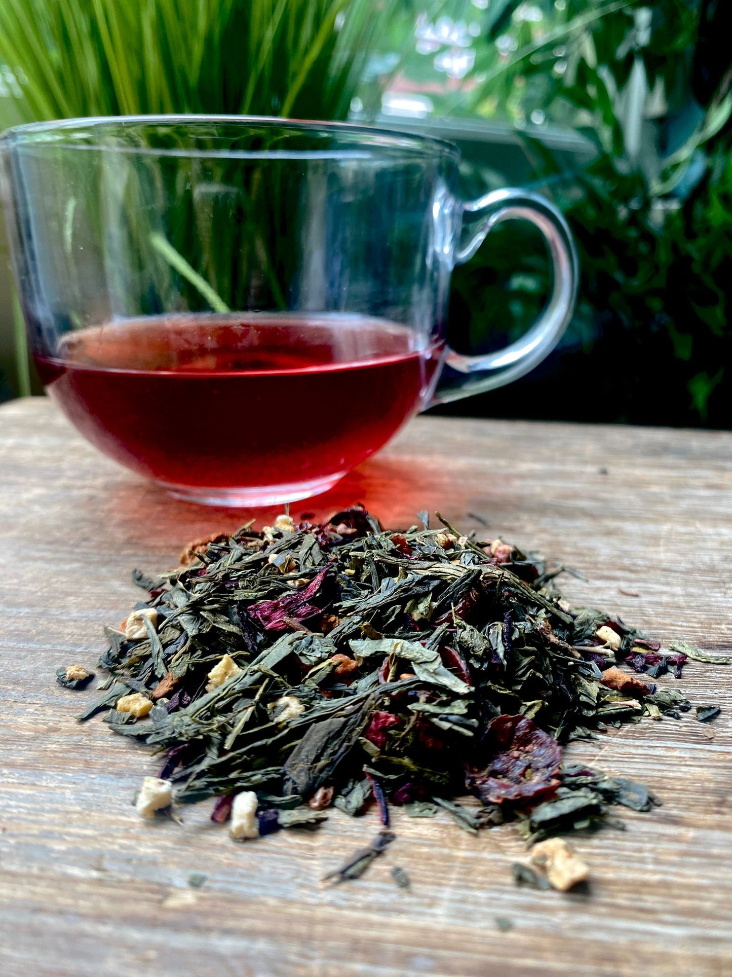 A glass cup of vibrant red tea sits on a wooden table surrounded by Peach and Berry Green Tea loose leaf tea leaves. Lush green plants are blurred in the background, creating a calming atmosphere.