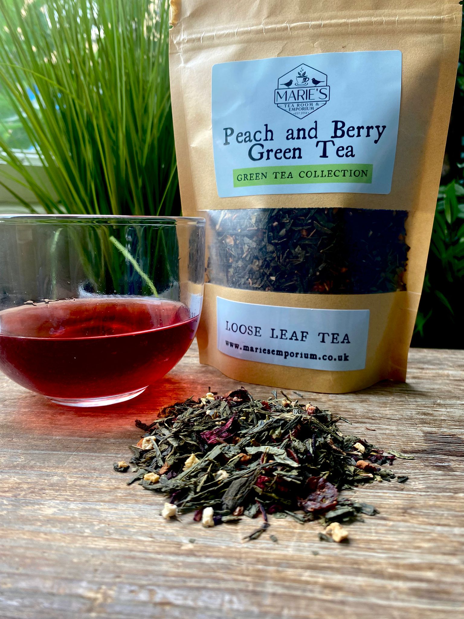 A glass of deep red tea sits on a wooden table alongside a pile of loose leaf tea. A maries peach and berry green tea package is in the background.