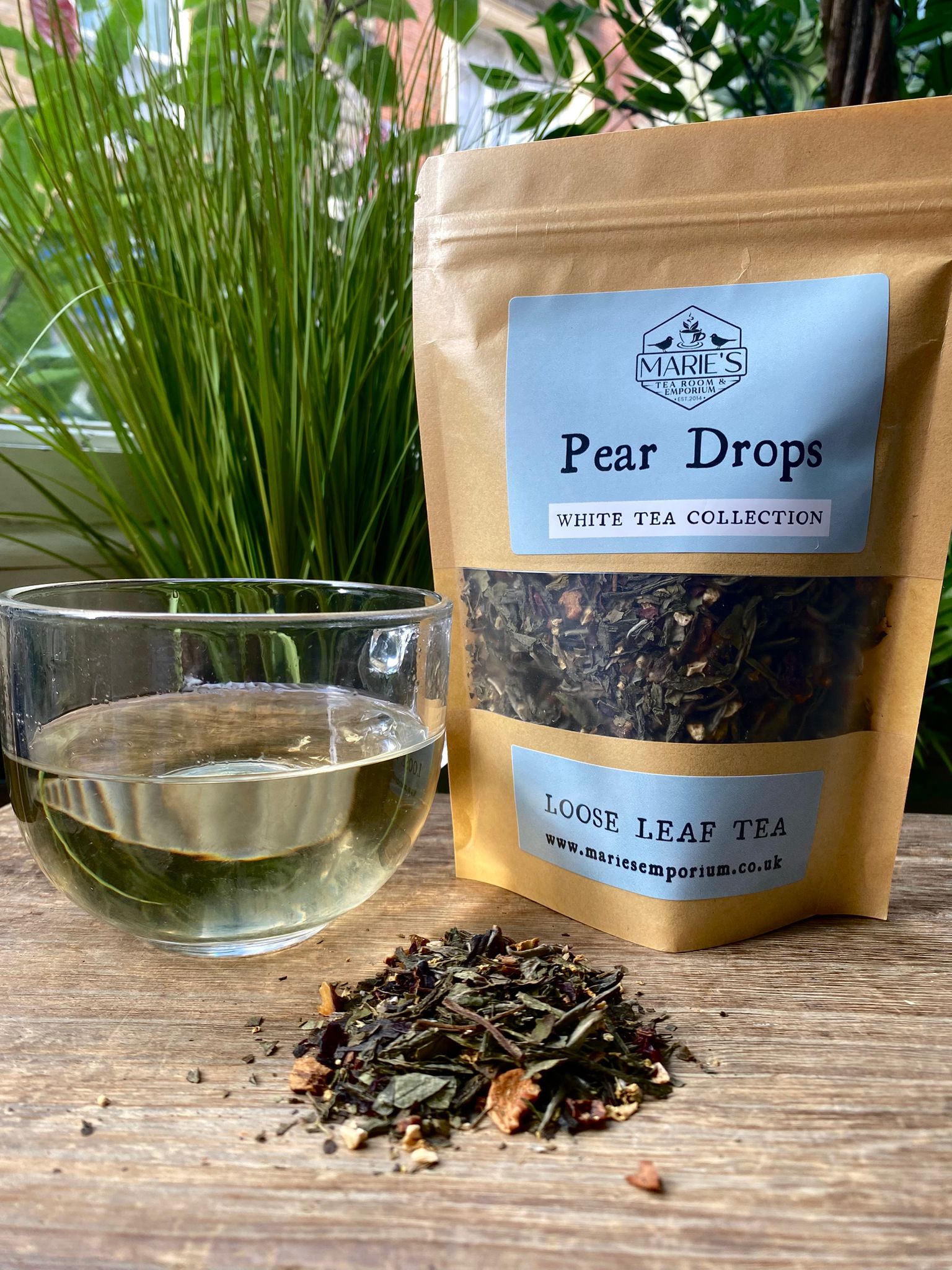 Bag of 'Pear Drops' white tea from Marie's Emporium with a glass of tea and loose tea leaves on a wooden surface.