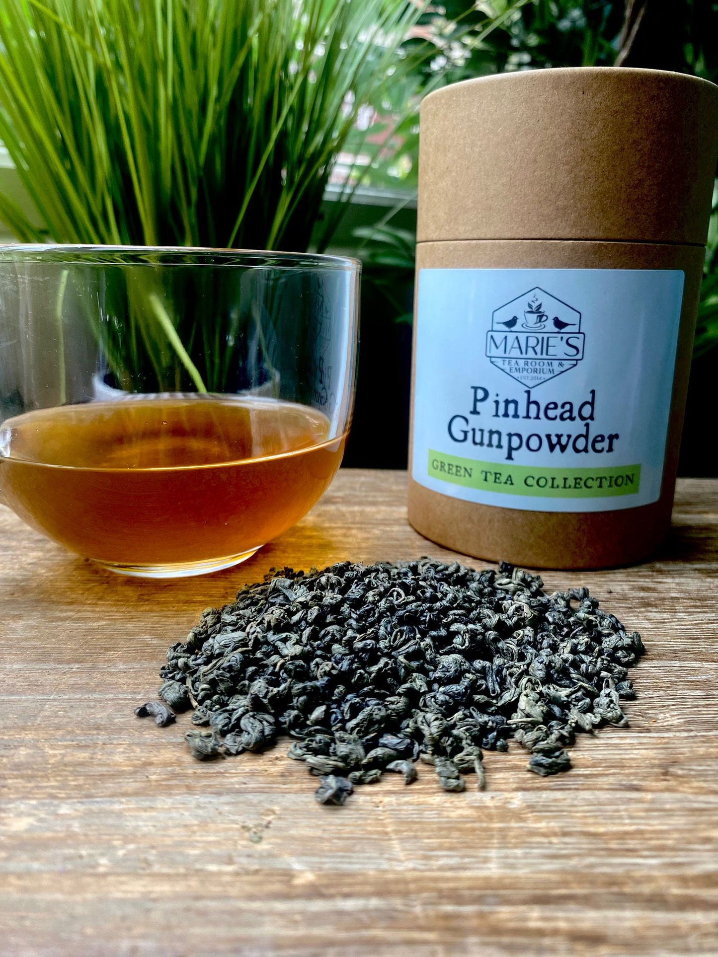maries Pinhead Gunpowder with tea leaves and a glass of tea on a wooden surface.