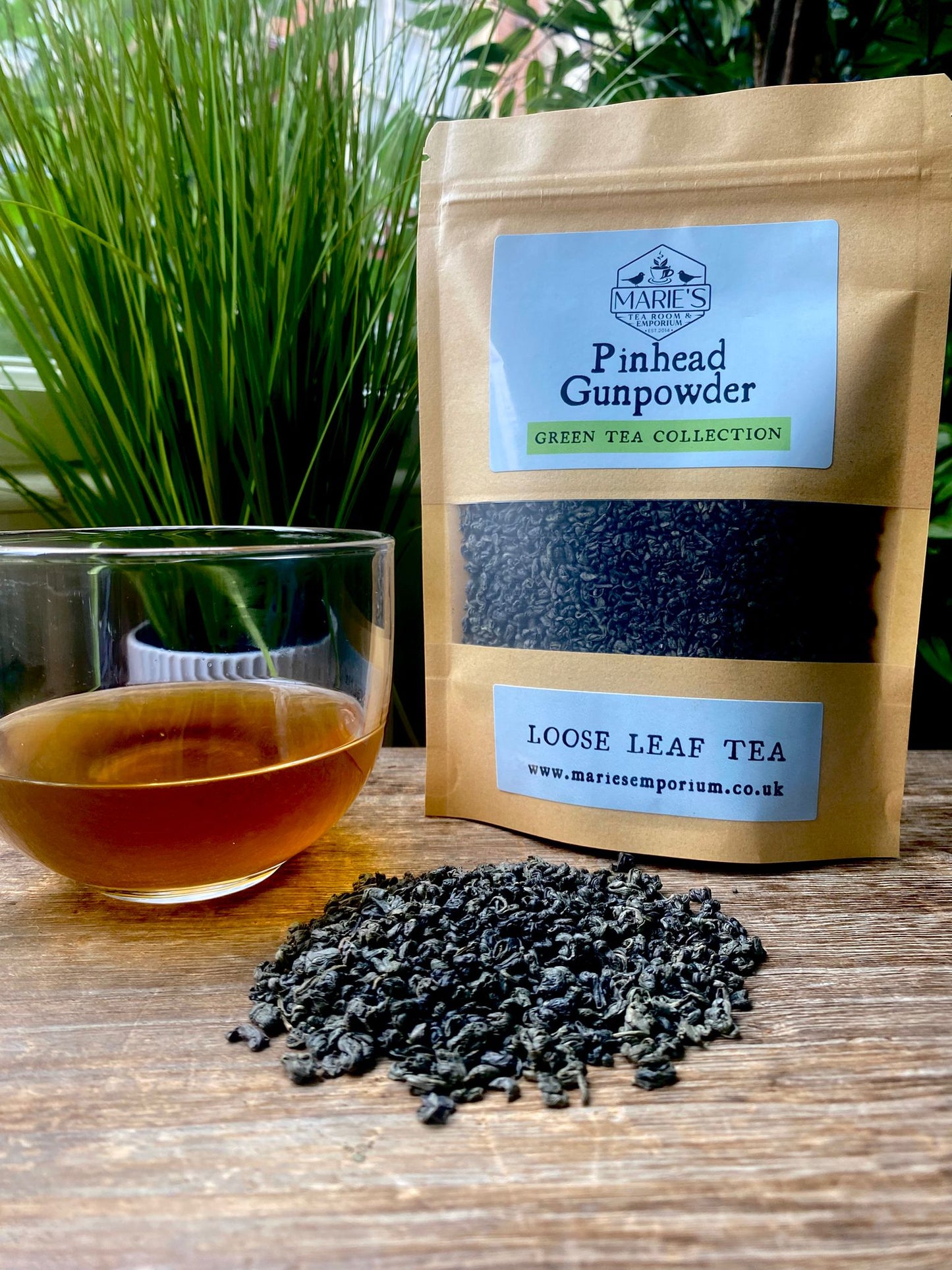 Bag of marie's Pinhead Gunpowder green tea with a glass of brewed tea and loose tea leaves on a wooden surface.