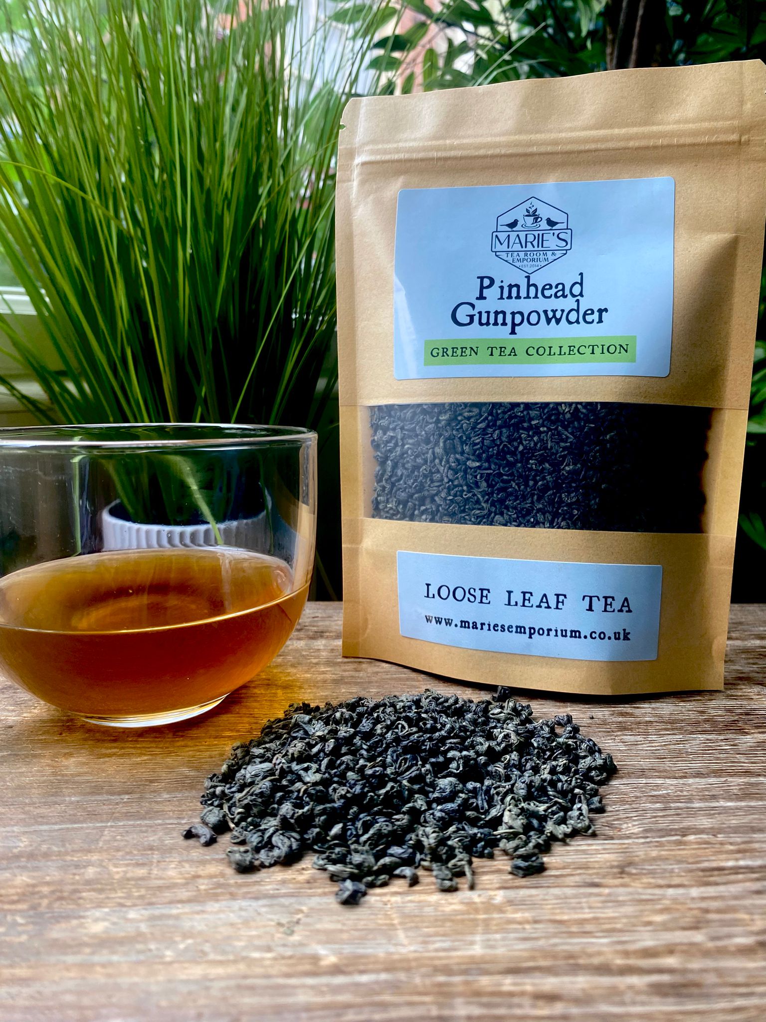 Bag of marie's Pinhead Gunpowder green tea with a glass of brewed tea and loose tea leaves on a wooden surface.