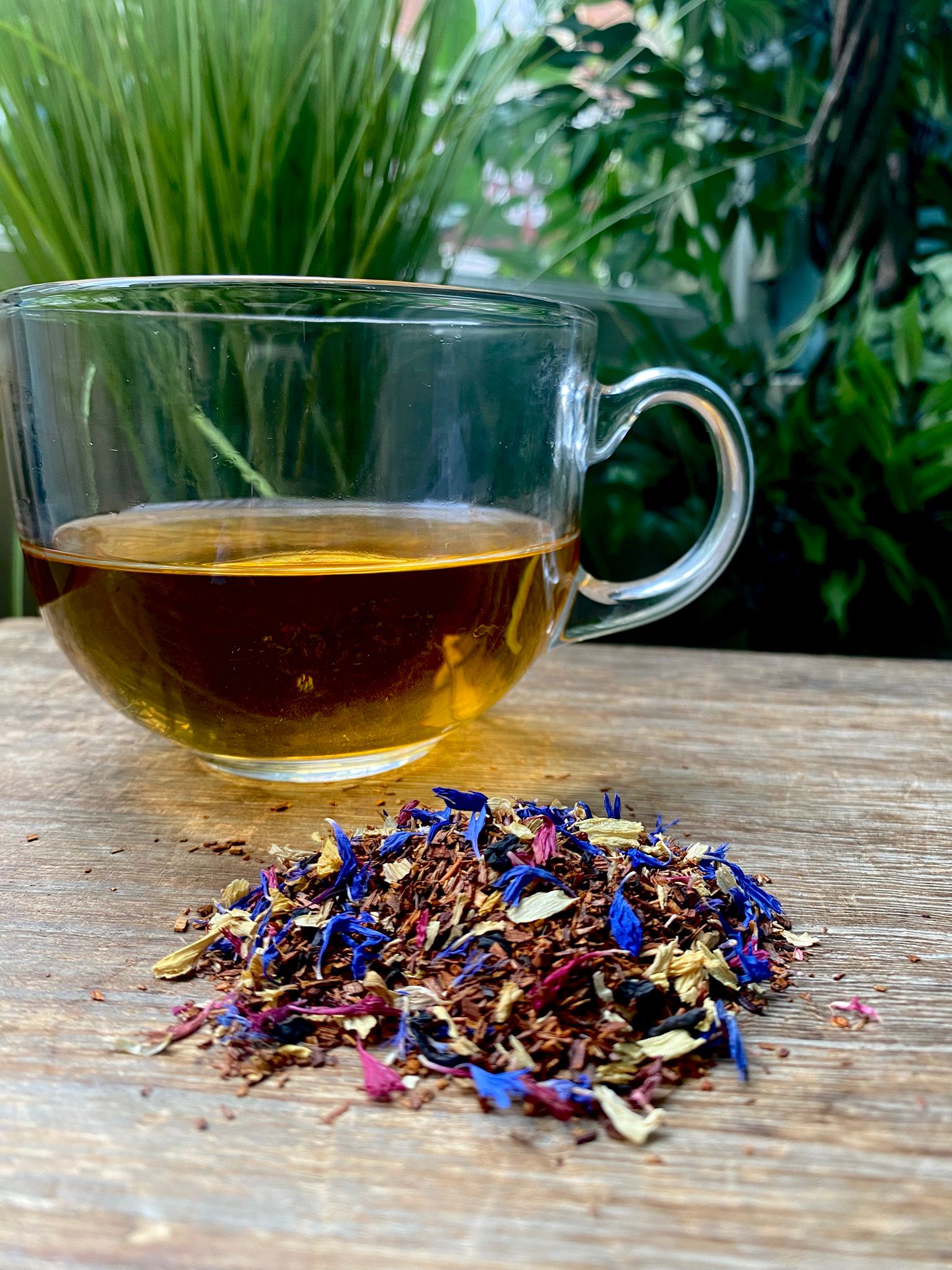 A clear glass cup filled with Marie's amber-colored Pocahontas herbal tea sits on a wooden surface. In front, a colorful pile of loose tea leaves and petals. Lush green foliage in background.