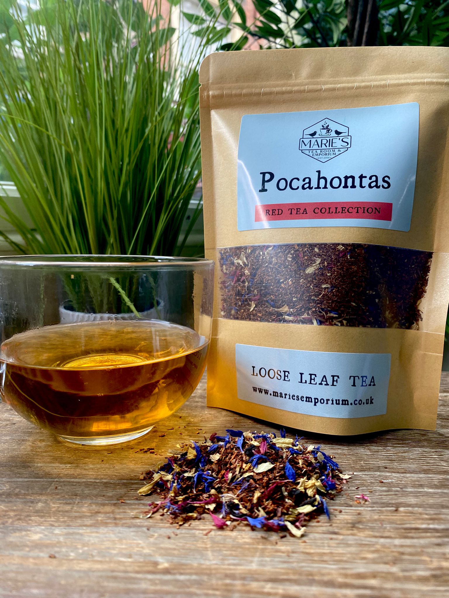 A glass cup of amber tea sits on a wooden table next to a pouch of maries Pocahontas loose leaf tea. Colorful tea leaves are scattered in front.