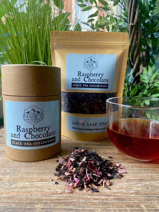 Maries Raspberry and chocolate packaging and loose tea leaves with a glass of tea on a wooden surface, surrounded by plants.
