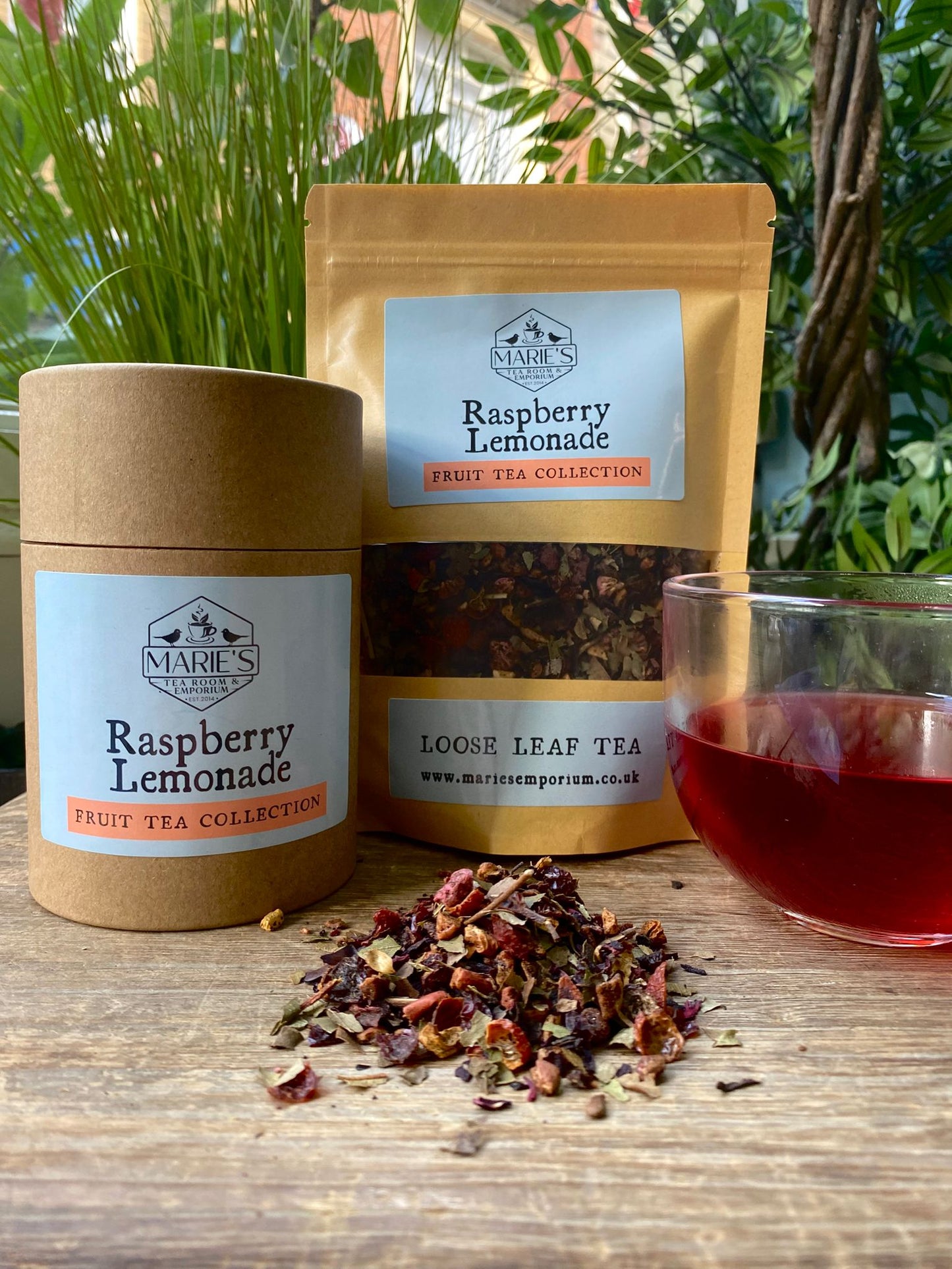 Marie's Emporium raspberry lemonade tea set with tea leaves, packaging, and a glass of tea on a wooden surface.