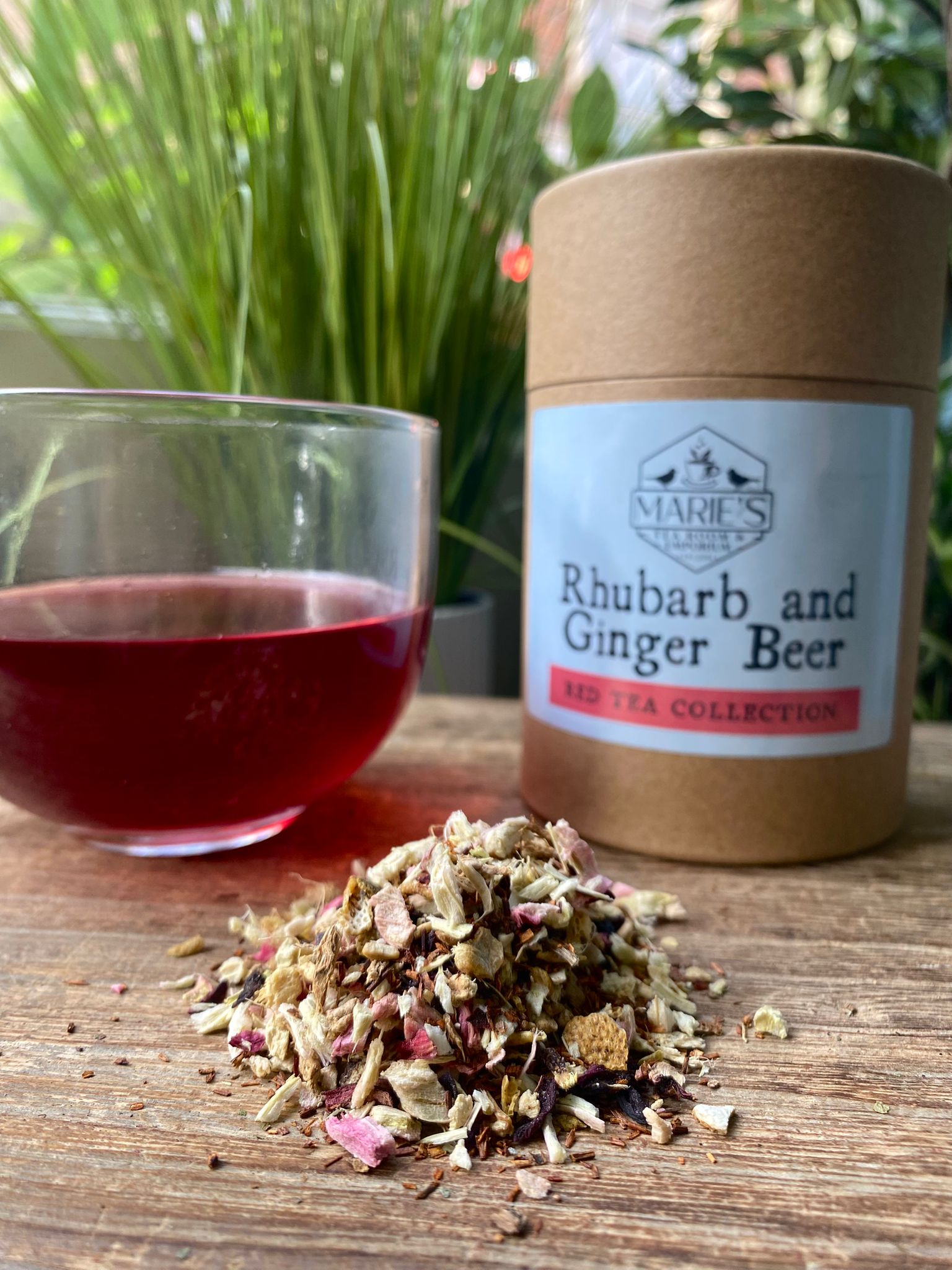 A glass of red tea beside a cylindrical container labeled "Marie's emporium rhubarb and ginger beer loose leaf tea " with loose leaves of the tea in front. Background of green foliage.
