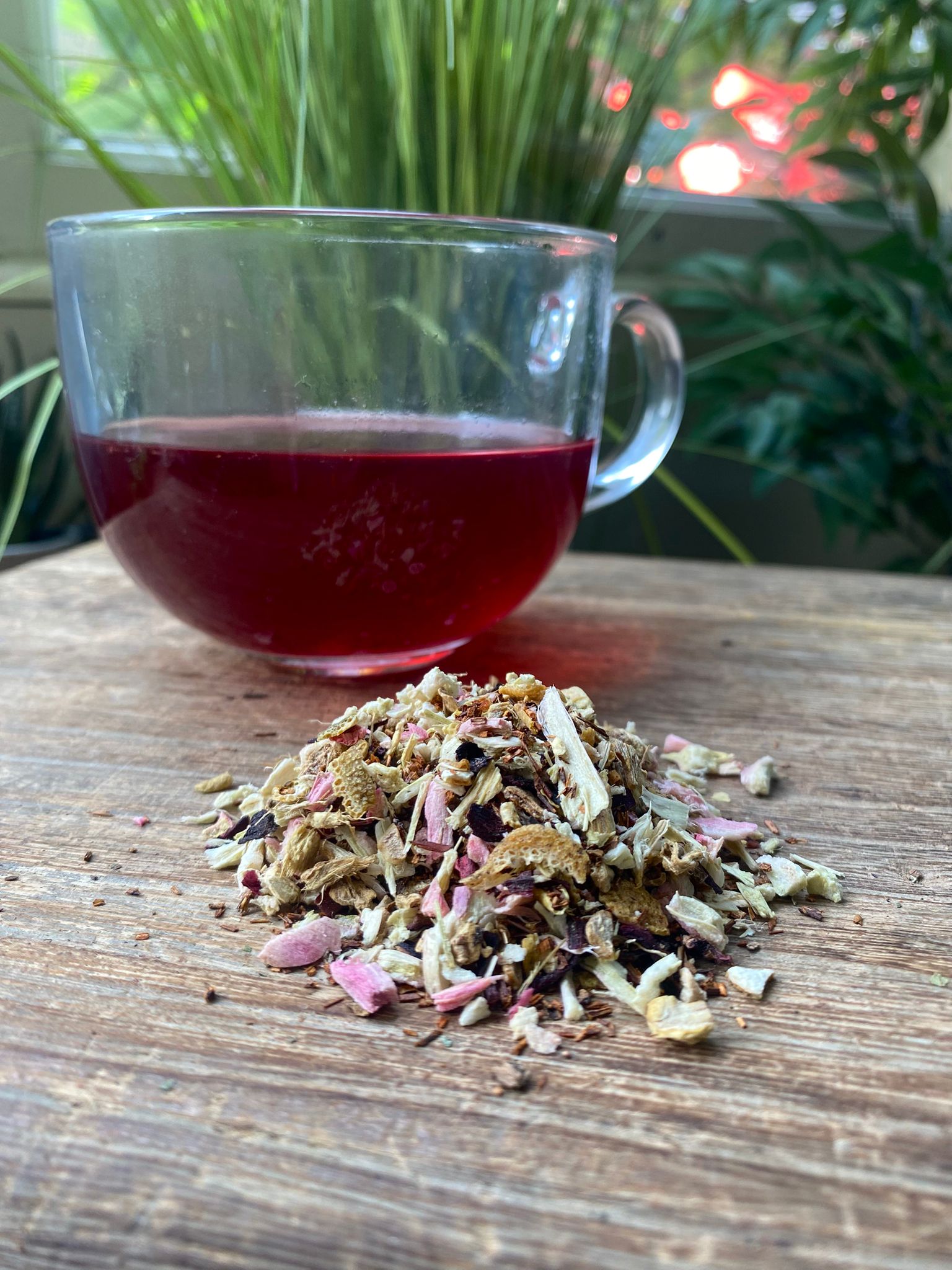 A pile of marie's rhubarb and ginger beer loose leaf tea sits on a wooden surface, with a clear cup of deep red tea behind it. Lush green plants are in the background.