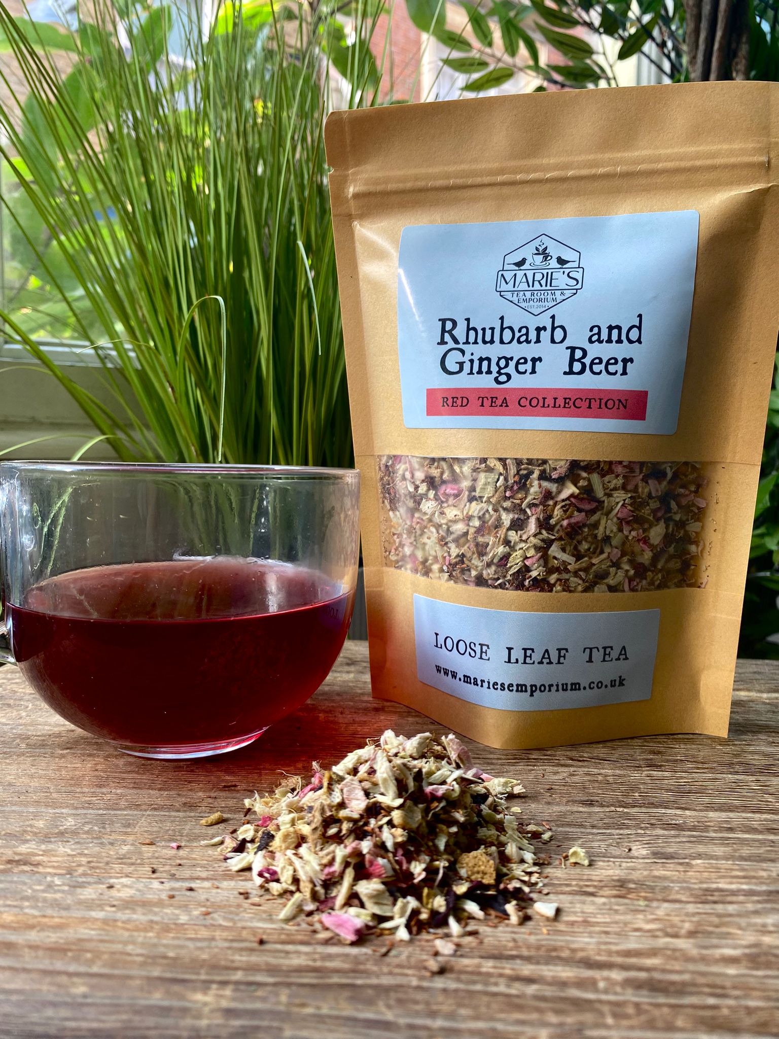 Clear glass cup of red tea beside a brown package labeled "Marie's rhubarb and ginger beer loose leaf tea " on a wooden table. Lush green plants in background.