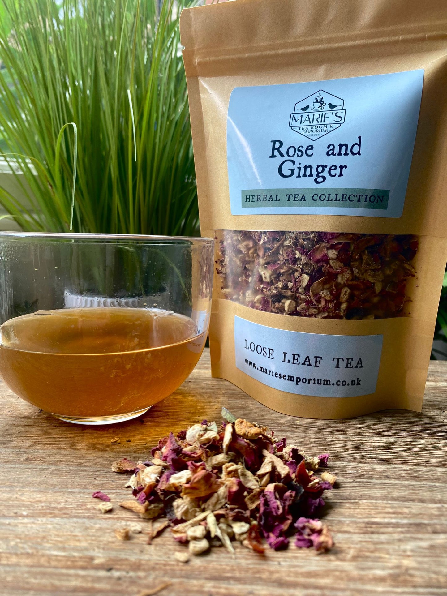 maries Bag of 'Rose and Ginger' herbal tea with a glass of brewed tea and scattered tea leaves on a wooden surface.