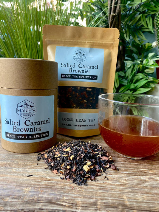 marie salted caramel brownie Tea packaging and loose tea leaves with a glass of tea on a wooden surface, surrounded by greenery.