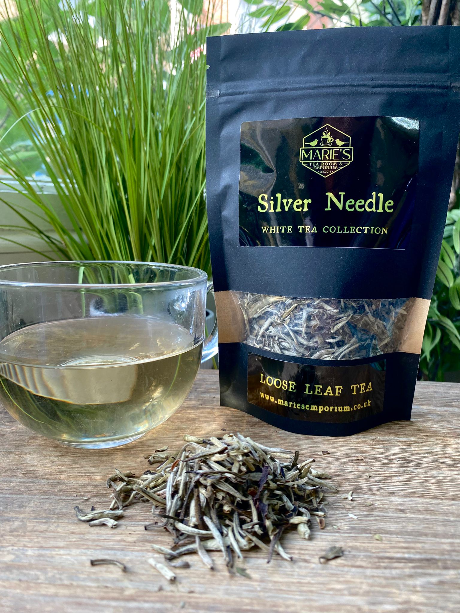 Marie's Emporium Silver Needle white tea package with a glass of tea and loose leaf tea on a wooden surface.