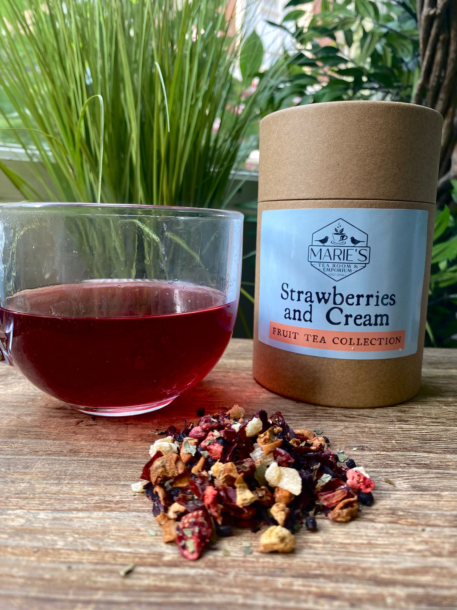 Tea canister labeled 'Strawberries and Cream' next to a glass of red tea and dried loose leaf tea leaves on a wooden surface with plants in the background