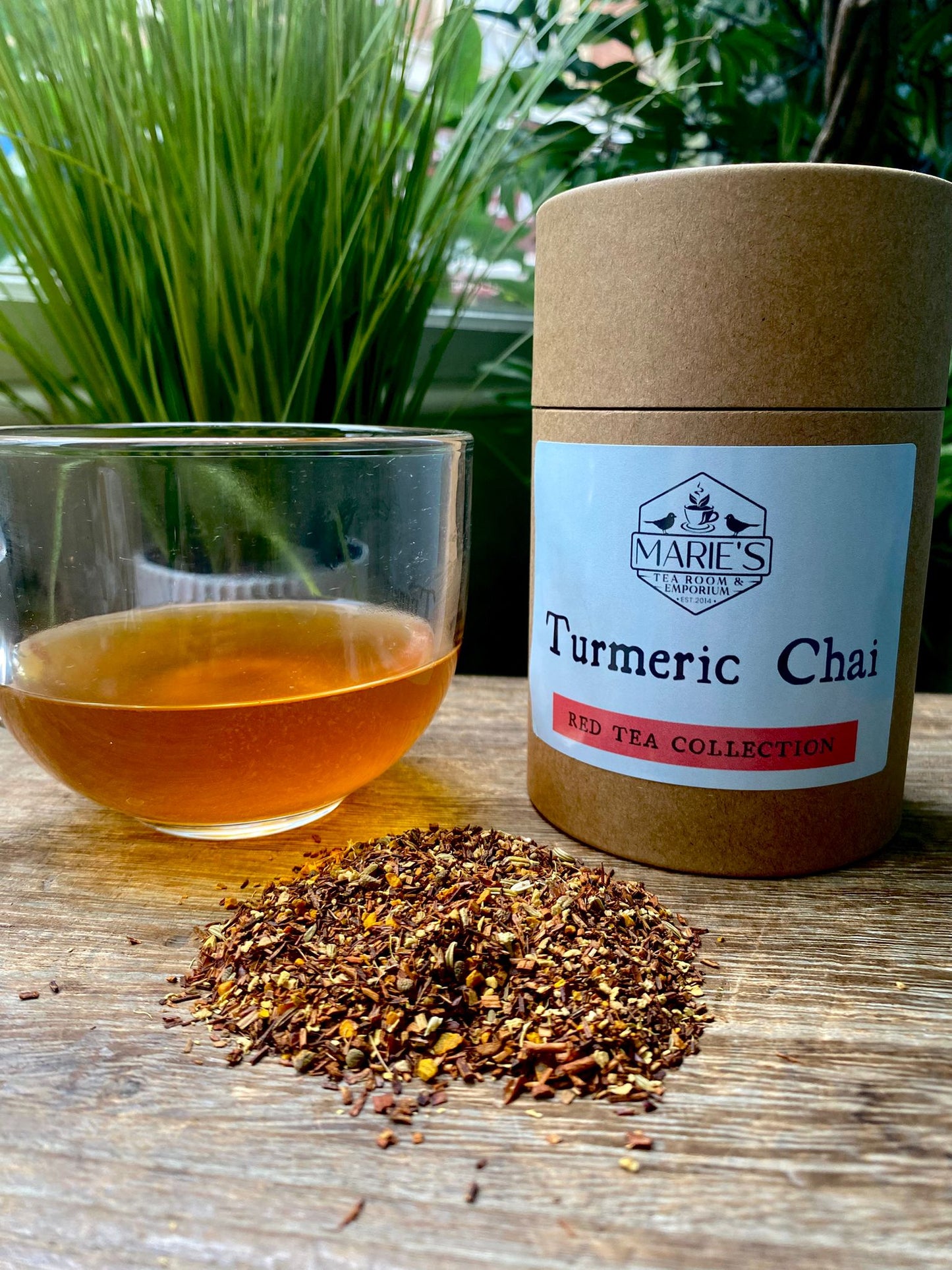 A glass cup of amber-colored turmeric chai tea sits beside a labeled canister and a small pile of loose tea leaves on a wooden table, with green plants in the background.