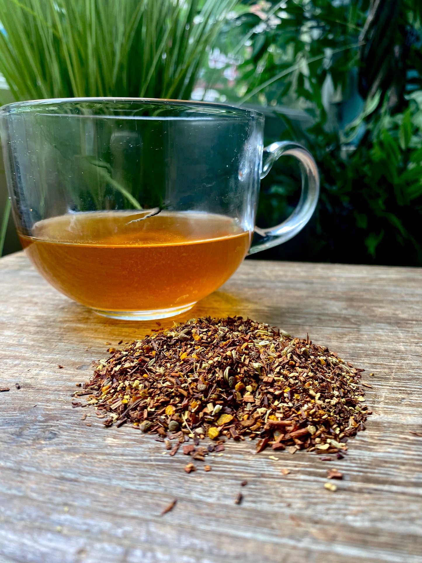 A clear glass cup filled with turmeric chai tea sits on a wooden surface beside a small pile of loose leaf tea. Lush green foliage is visible in the background.