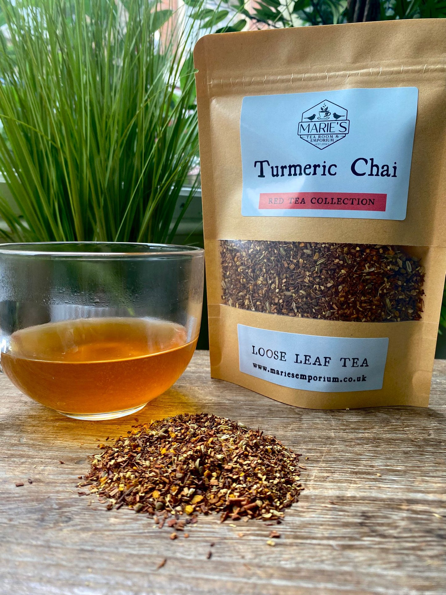 A clear glass cup with amber-colored turmeric chai sits beside a brown paper bag labeled "Turmeric Chai Loose Leaf Tea," with a pile of tea leaves on a wooden surface. The background features lush green plants, creating a fresh and natural ambiance.