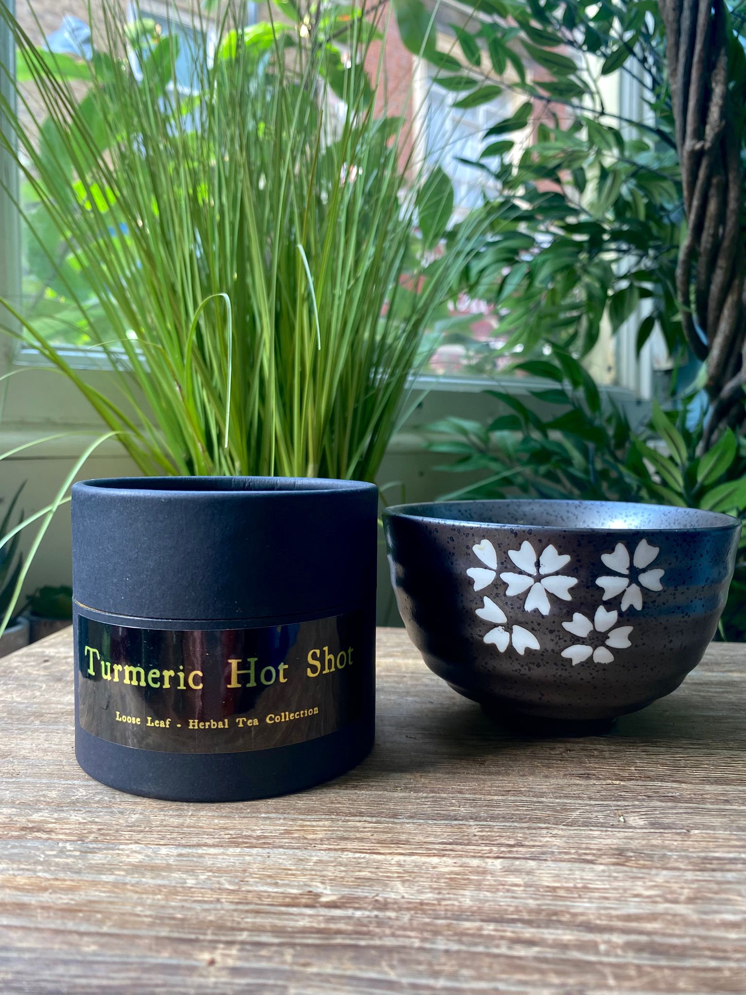 Marie's Turmeric hot shot container  containing loose leaf tea next to a serving bowl on a wooden surface with plants in the background.