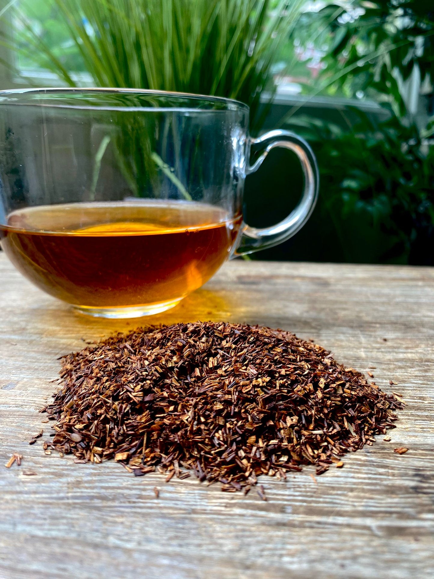Clear glass cup of vanilla rooibos loose leaf tea with tea leaves on a wooden surface with greenery in the background