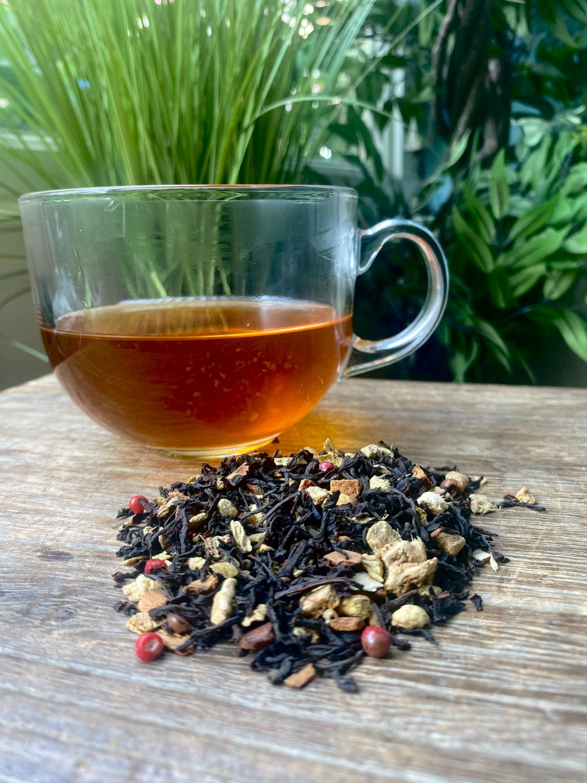 Clear glass mug with Vanilla Spice black tea maries collection tea and a pile of loose tea leaves on a wooden surface with plants in the background