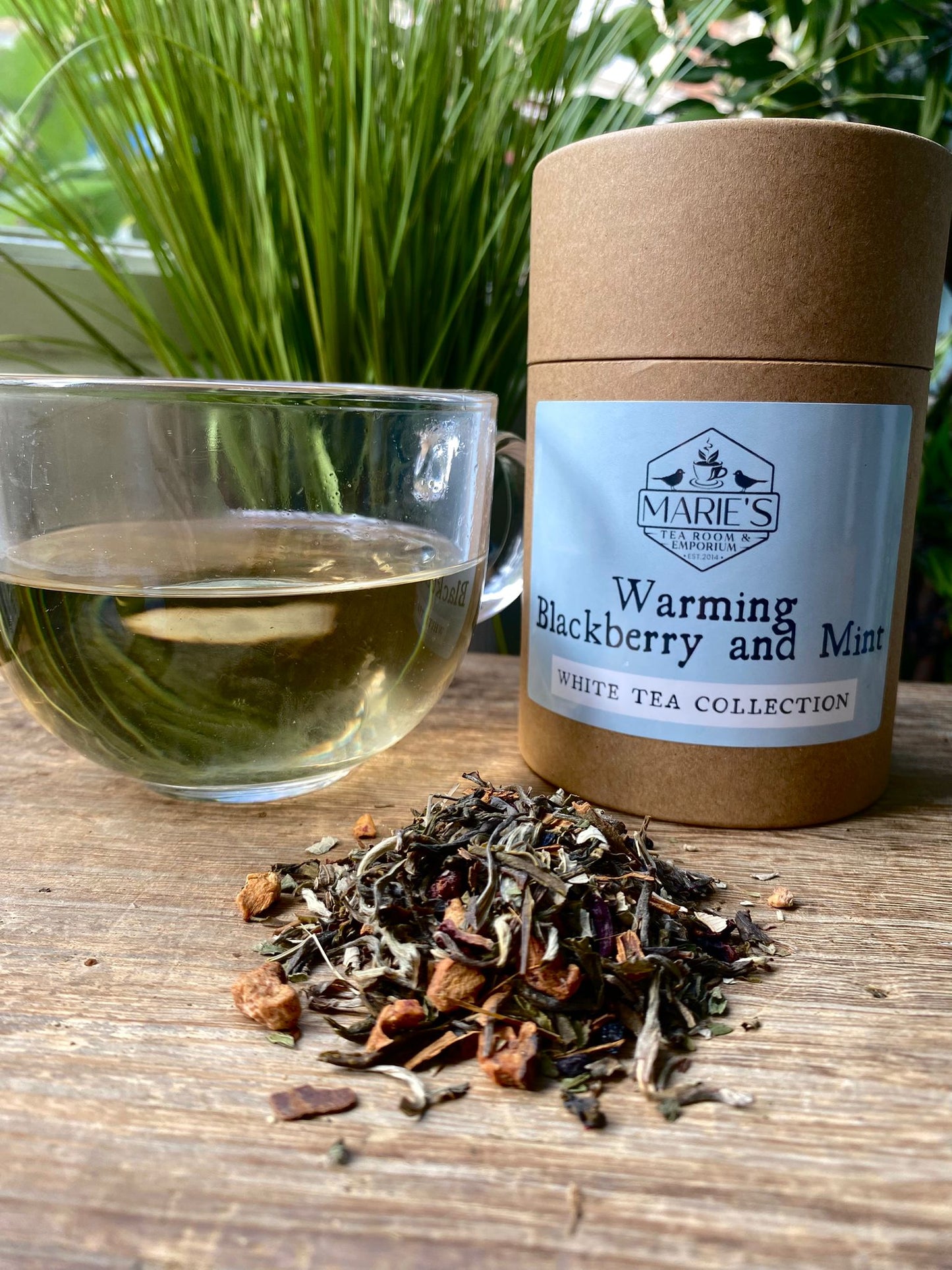 Marie's Emporium warming blackberry and mint tea packaging with loose leaf tea leaves and a glass of tea on a wooden surface.
