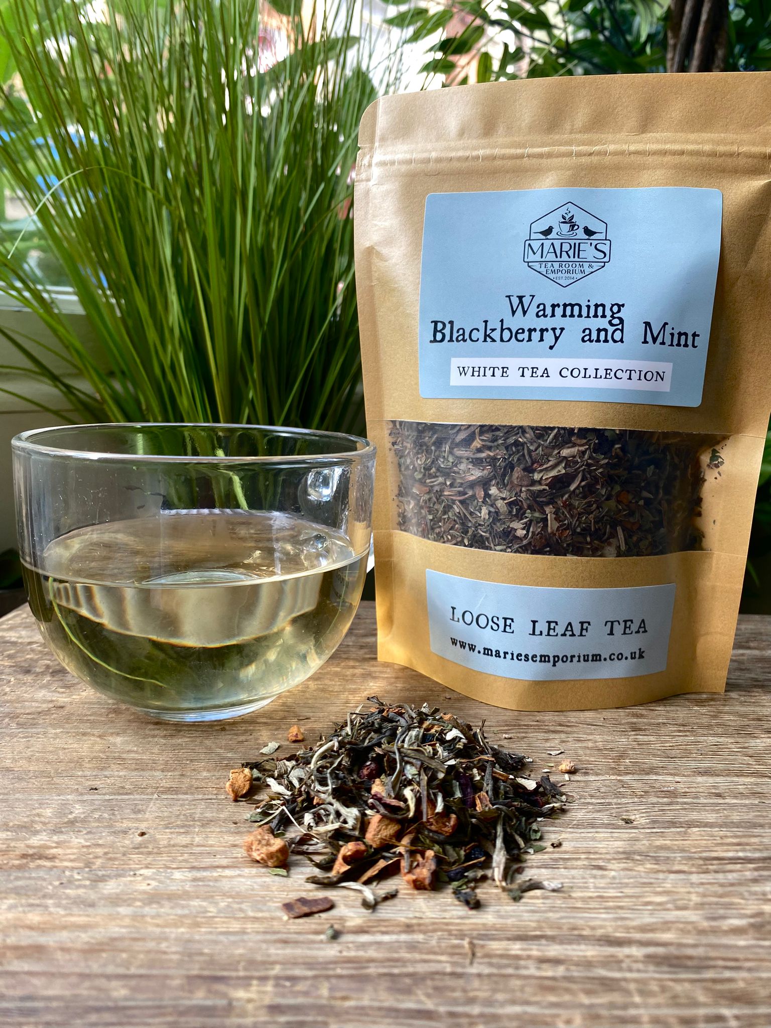Marie's Emporium warming blackberry and mint tea package with a glass of brewed tea and loose leaf tea leaves on a wooden surface.