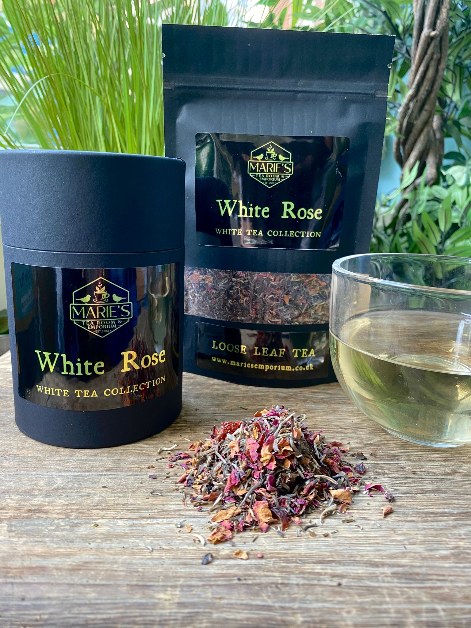Maries White Rose tea packaging with a black canister and pouch, alongside a pile of loose tea leaves. A glass cup filled with brewed tea. Greenery in the background.