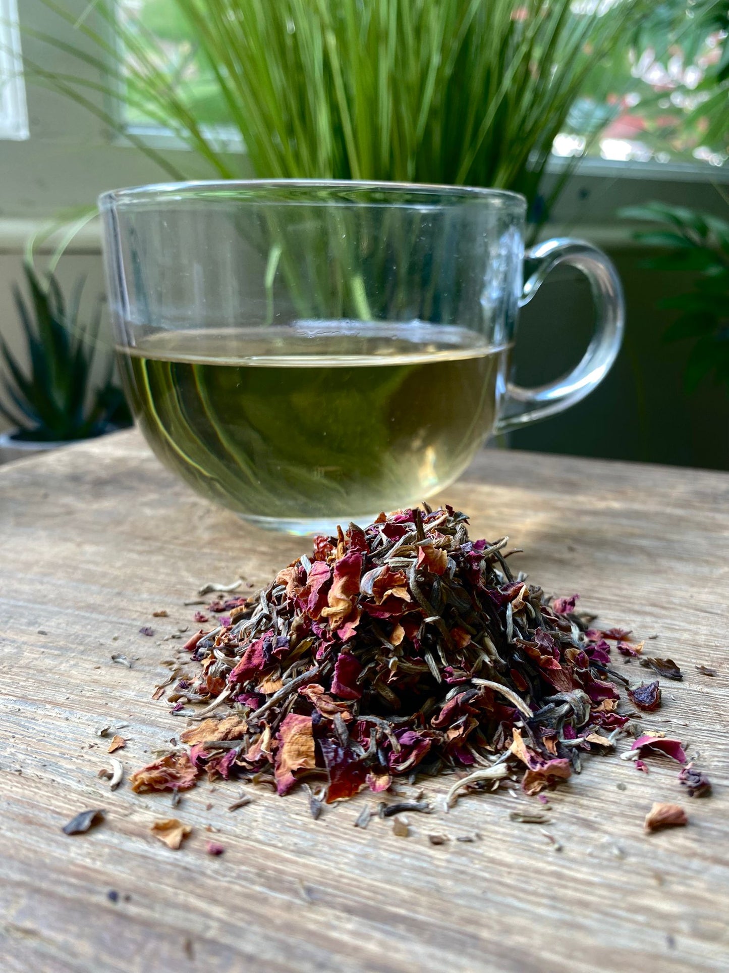 A glass mug of herbal tea sits on a wooden table, with a pile of maries white rose tea in the foreground. Greenery is visible in the blurred background