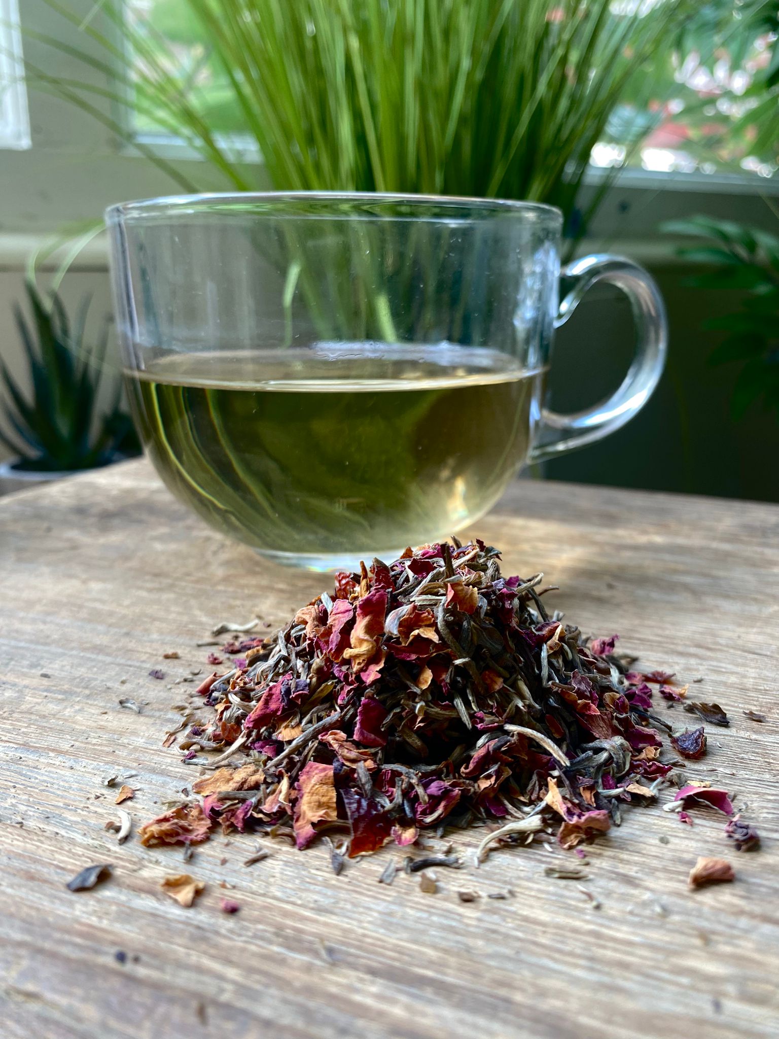 A glass mug of herbal tea sits on a wooden table, with a pile of maries white rose tea in the foreground. Greenery is visible in the blurred background